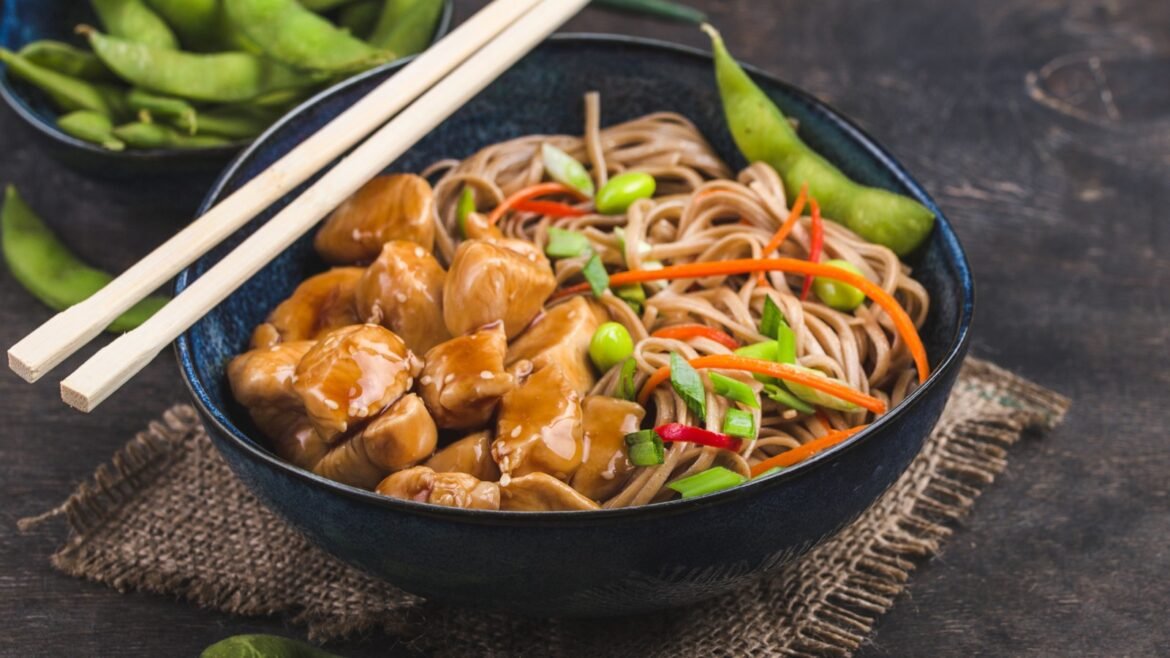 Bowl of saucy chicken and soba noodles with carrots, edamame and green onions, with chopsticks resting on the bowl rim.