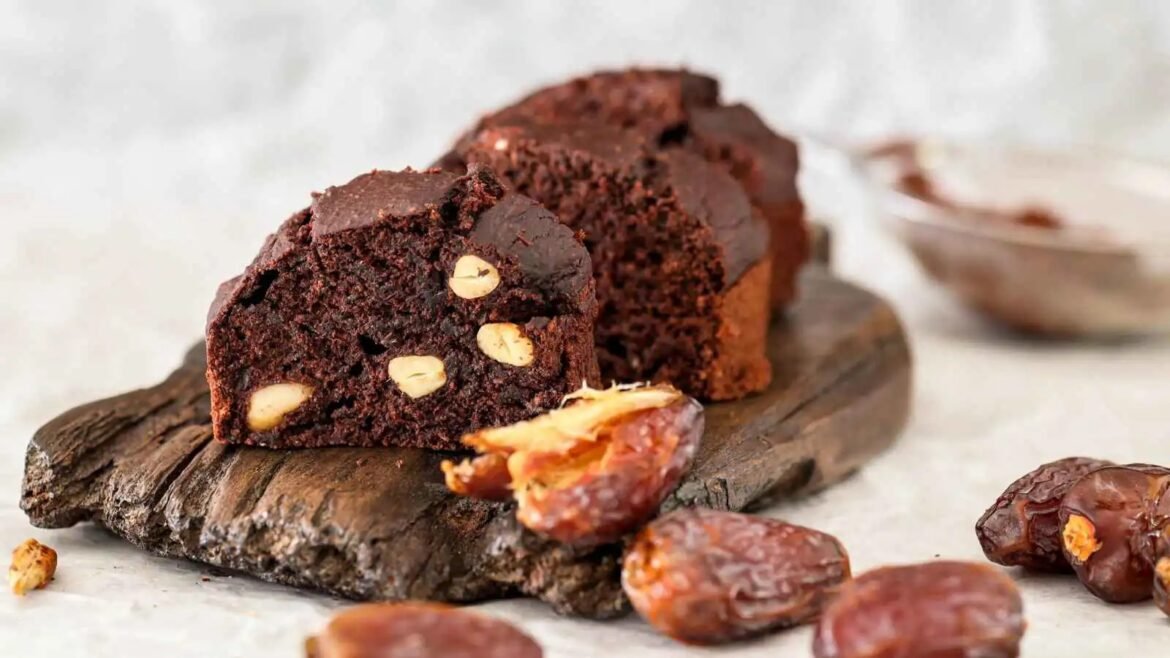Chunky chocolate brownie loaf with almonds on a wooden board, with date pieces scattered around.