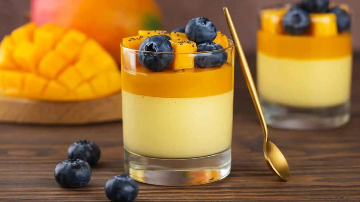 Two-layer mango pudding in a glass topped with blueberries and mango cubes, with a gold spoon beside it on a wooden table.