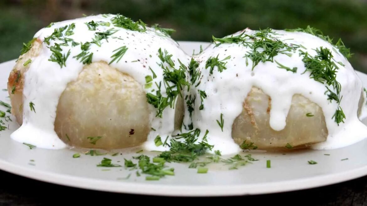 Two dumplings covered in white sauce and chopped dill on a white plate, served outdoors.