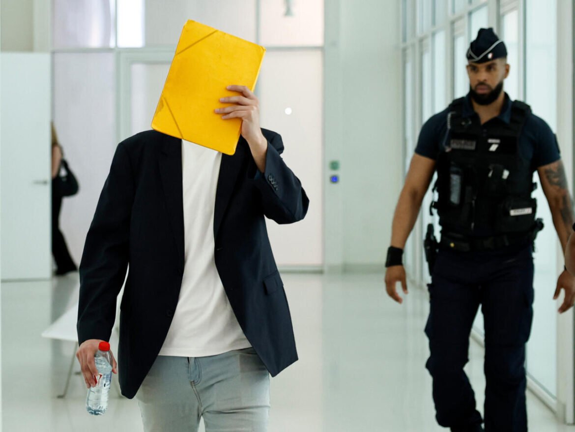 Man in a blazer hides his face behind a large yellow folder, holding a water bottle as an armed security officer follows in a bright hallway nearby.