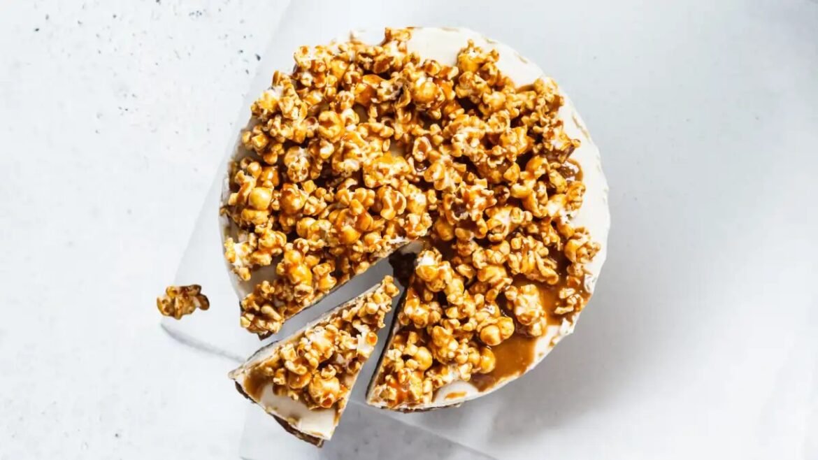 Round cake with white frosting topped generously with caramel popcorn, a slice removed to reveal the interior.