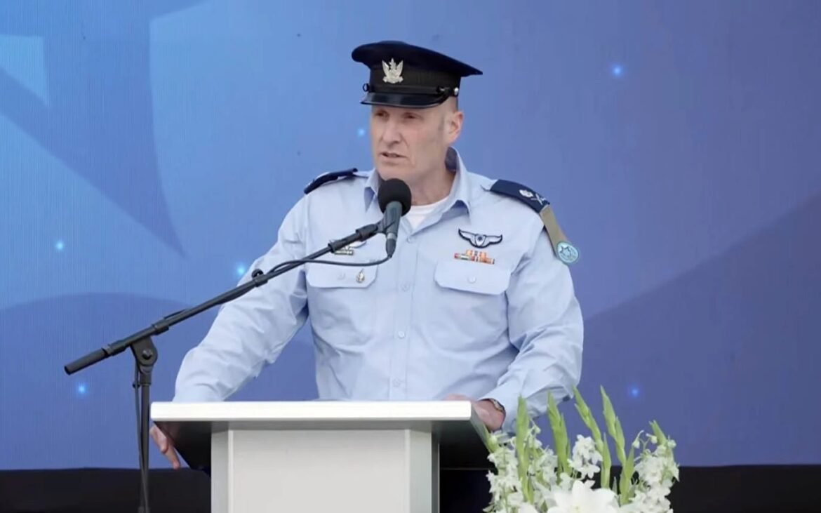 Uniformed officer at a podium delivering a speech, microphone in front, blue backdrop and white flowers nearby.