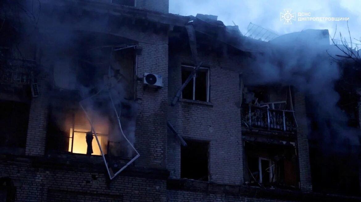 A brick apartment building on fire with thick smoke and orange flames visible through a window, a silhouette standing on a burning floor.