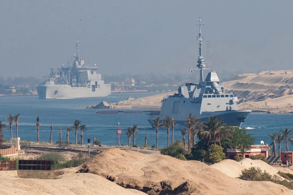 Two large gray warships anchored near a sandy coastline with palm trees in the foreground.