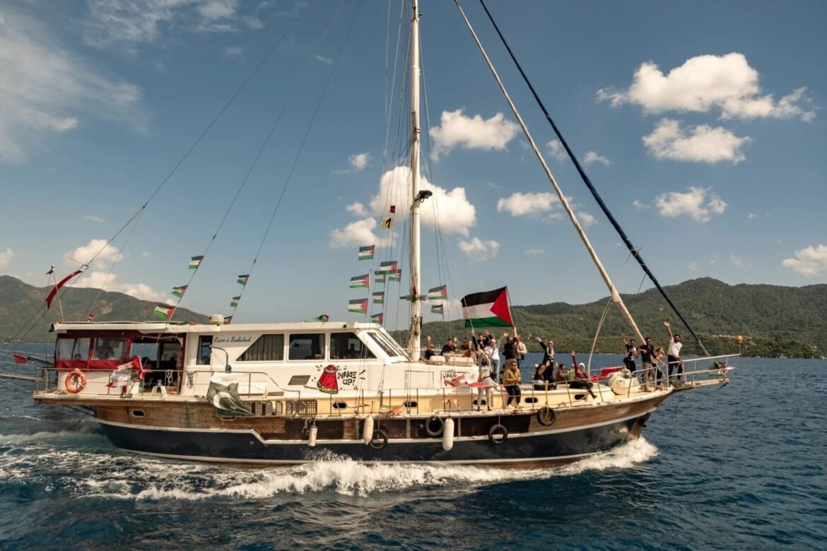 A white catamaran yacht with numerous people on the deck waving, flags strung along the rigging, sailing on blue water with a hilly coastline in the background under a clear sky.