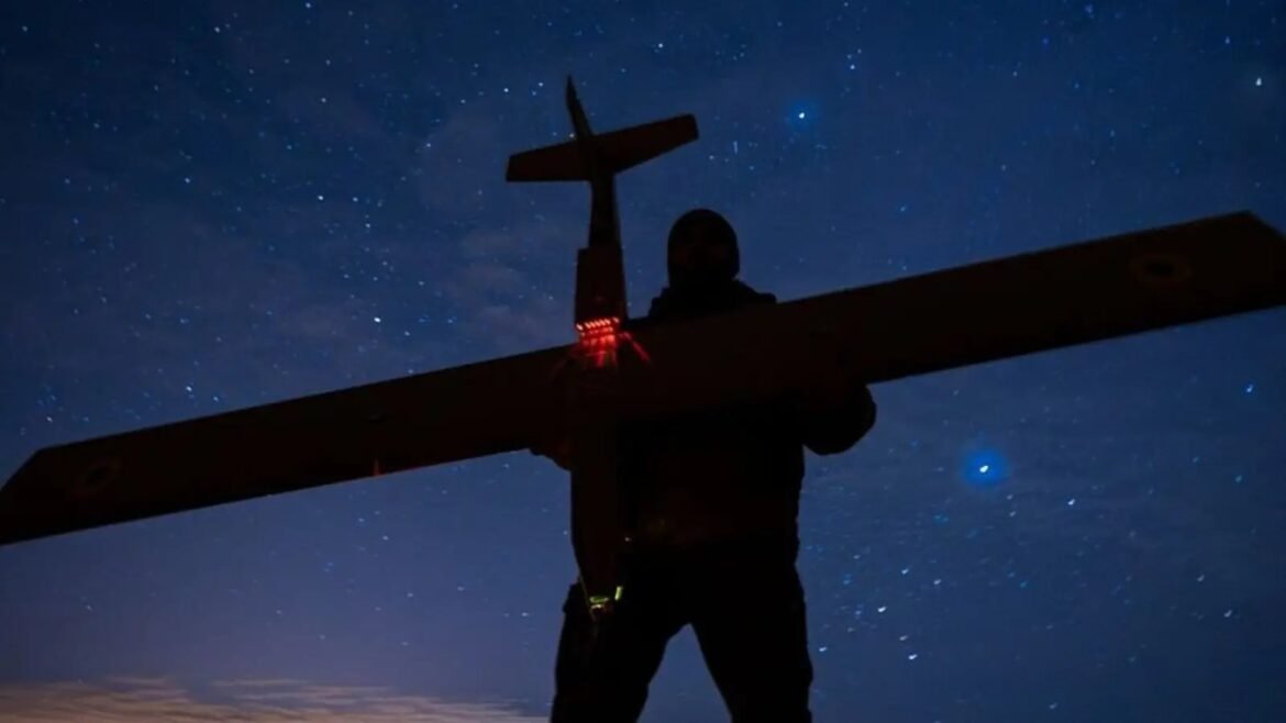 Silhouette of a person holding a large cross-shaped wing or frame with red lights, set against a starry night sky and horizon glow.