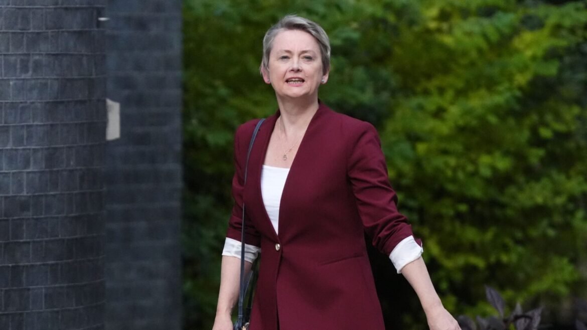 Professional woman with short gray hair wearing a burgundy blazer and white top, walking outdoors beside a brick structure and greenery in the background, carrying a bag strap.