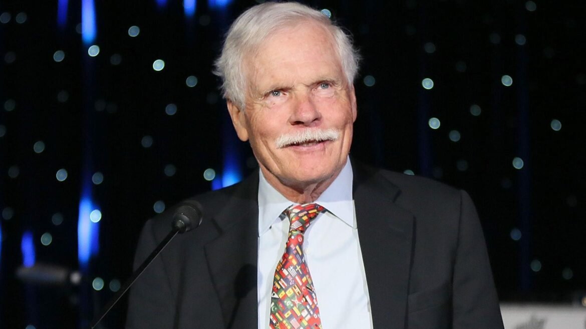 Senior man with white hair and mustache in a suit speaking into a microphone on a stage with blue, bokeh lights in the background.