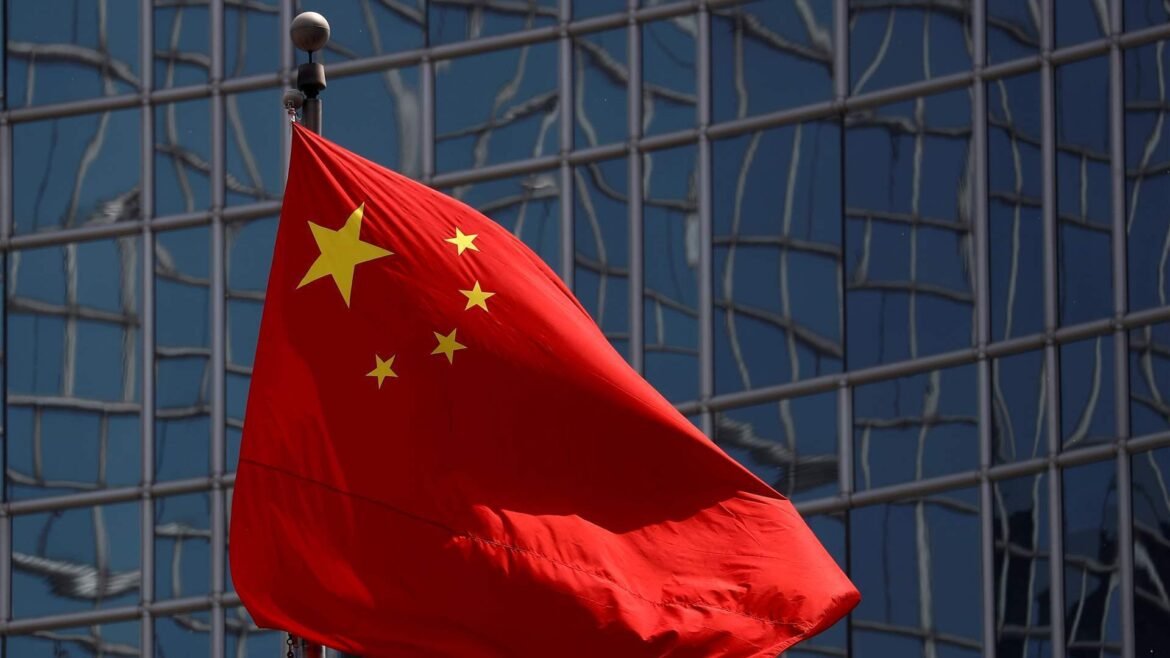Red Chinese flag with five yellow stars waving in front of a reflective glass building facade