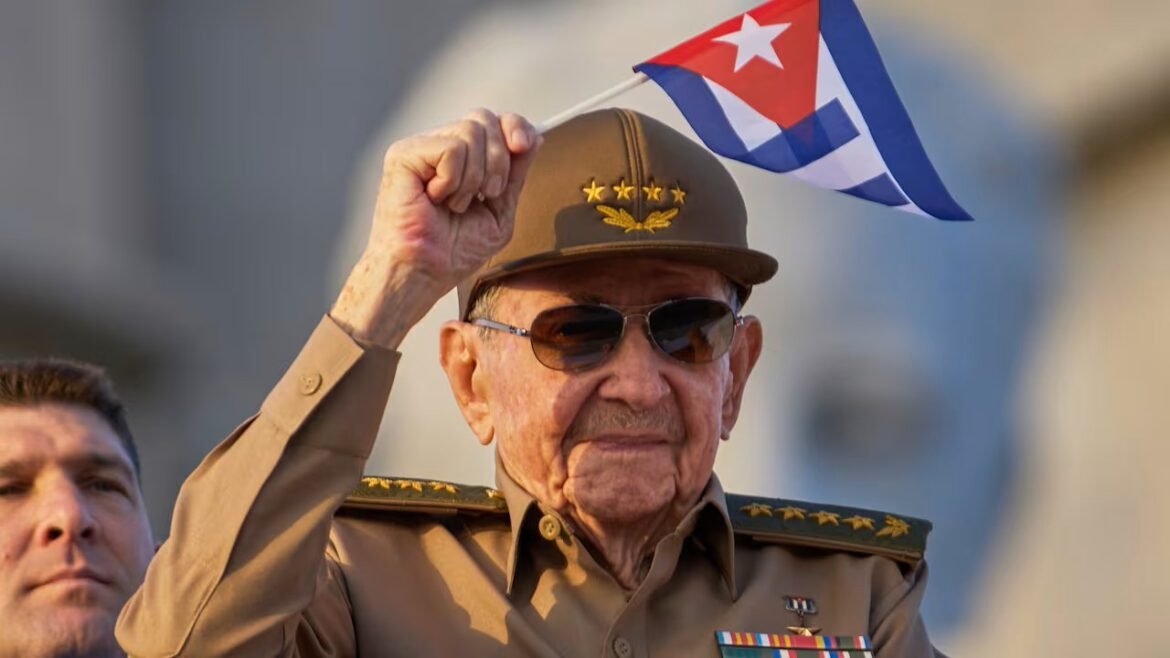 Elderly veteran in a tan military uniform waves a Cuban flag during a parade, wearing sunglasses and a cap with stars.