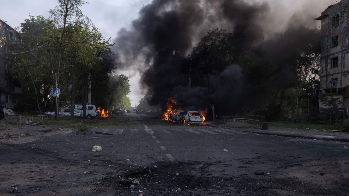 Cars on fire along a city street with thick black smoke rising above trees and surrounding buildings, debris on the road