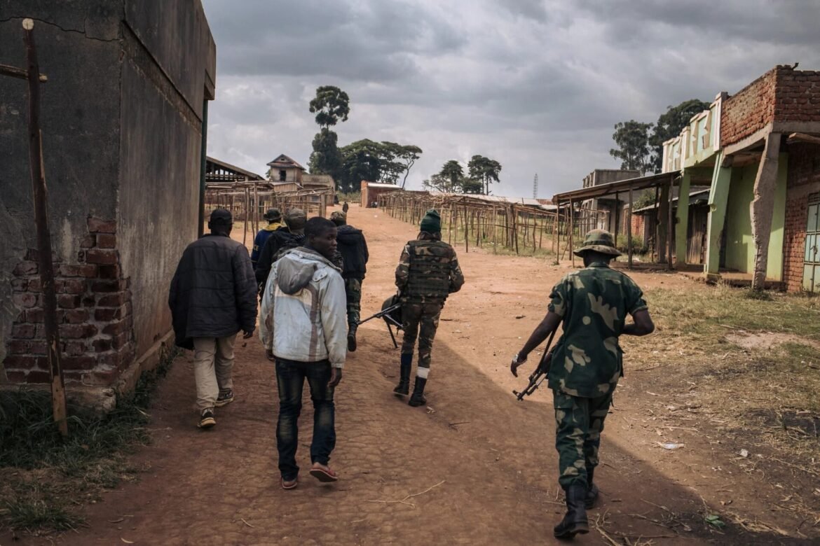 A squad of soldiers patrolling a dusty road between damaged brick buildings in a war-torn town under a cloudy sky.