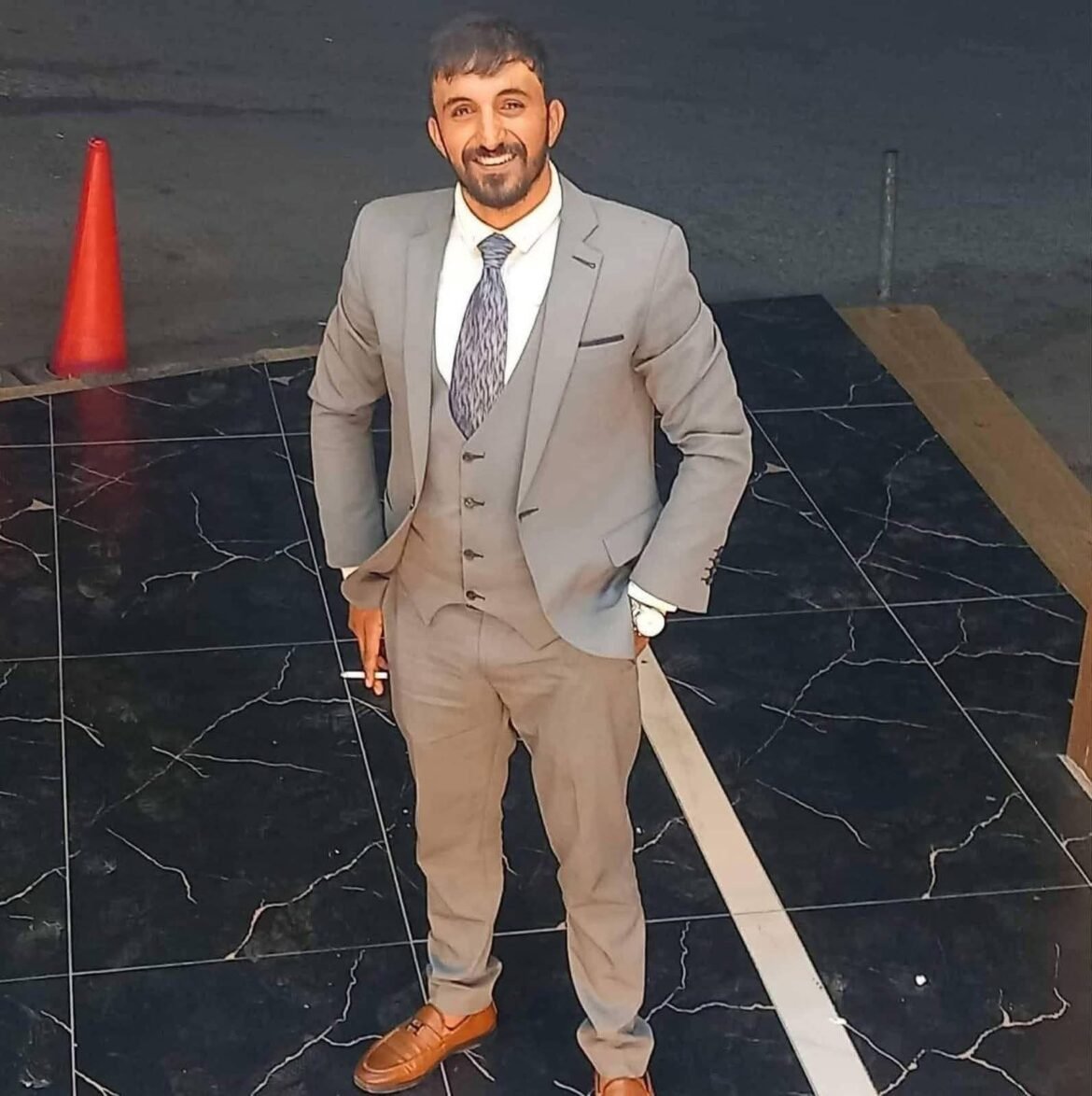 Man in a light gray suit and patterned tie standing on a black marble floor, smiling at the camera with an orange cone in the background.