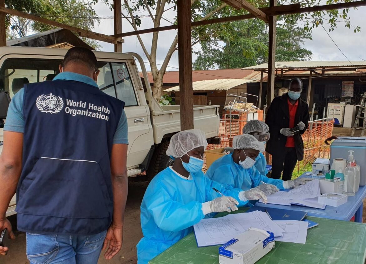 WHO workers in blue protective gowns and masks sit at a table signing forms outdoors, with a white pickup truck nearby.