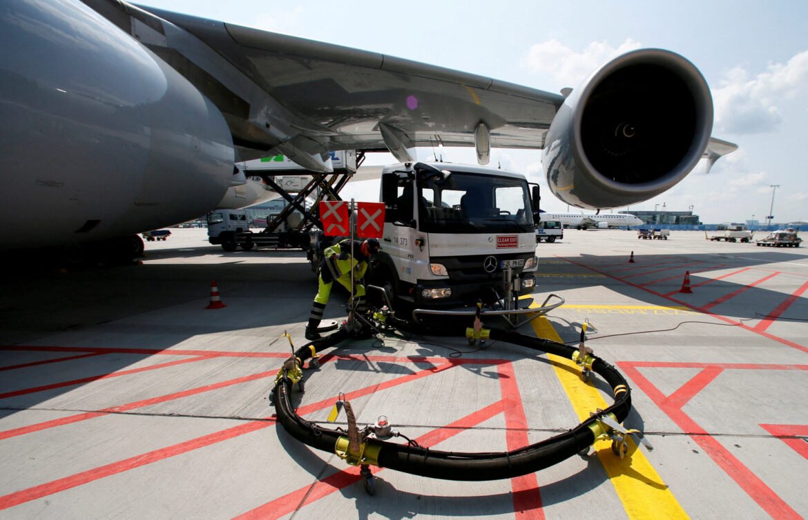 Ground crew services a large passenger jet on the tarmac with hoses and equipment under the wing.