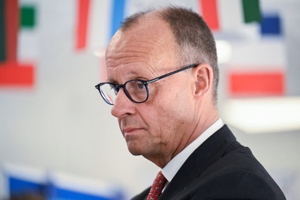 Side profile of a middle-aged man wearing a dark suit and red tie, with round blue glasses, in an office setting.