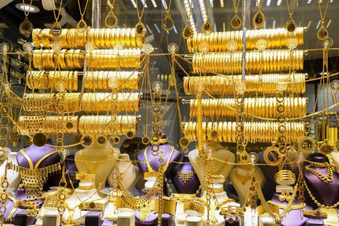 Gold jewelry display in a shop window with bracelets and necklaces arranged on purple busts behind glass.