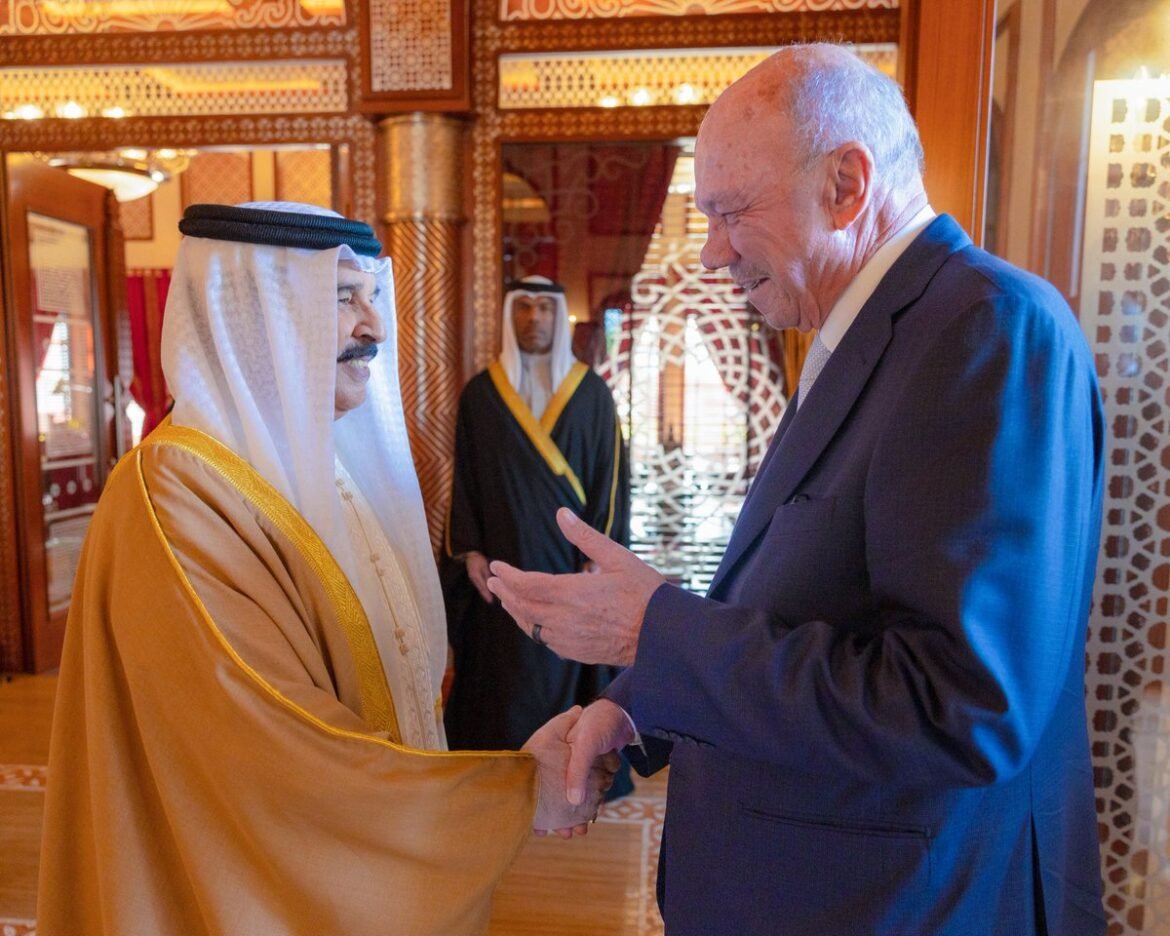 Two men shake hands in a richly decorated room; one wears a white headscarf and gold-trimmed robe, the other a dark suit.