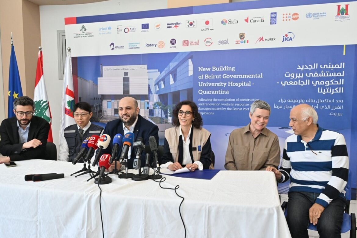 Press conference with six speakers seated at a white-clothed table, multiple microphones, and a blue project banner in the background showing Beirut Hospital construction details. Flags line the left side of the scene.