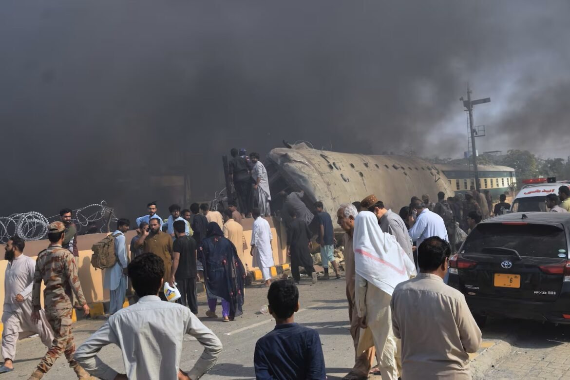 Crowd gathers around a crashed, burning train car on a road, thick smoke in the air and barbed wire beside the scene.