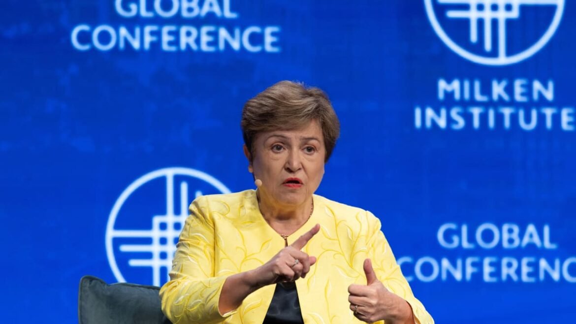 Woman in a yellow blazer speaking on stage at the Milken Institute Global Conference, with a blue backdrop and logos nearby.