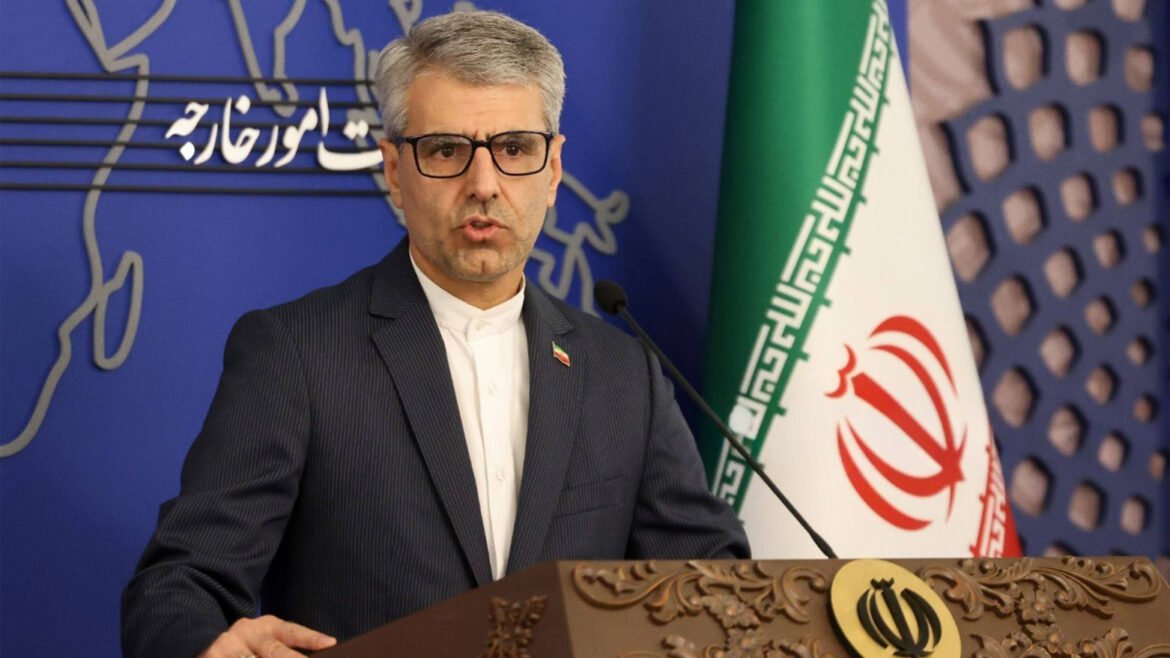 Man in a dark suit and glasses delivering a speech at a podium, with the Iranian flag behind him and a blue backdrop behind.