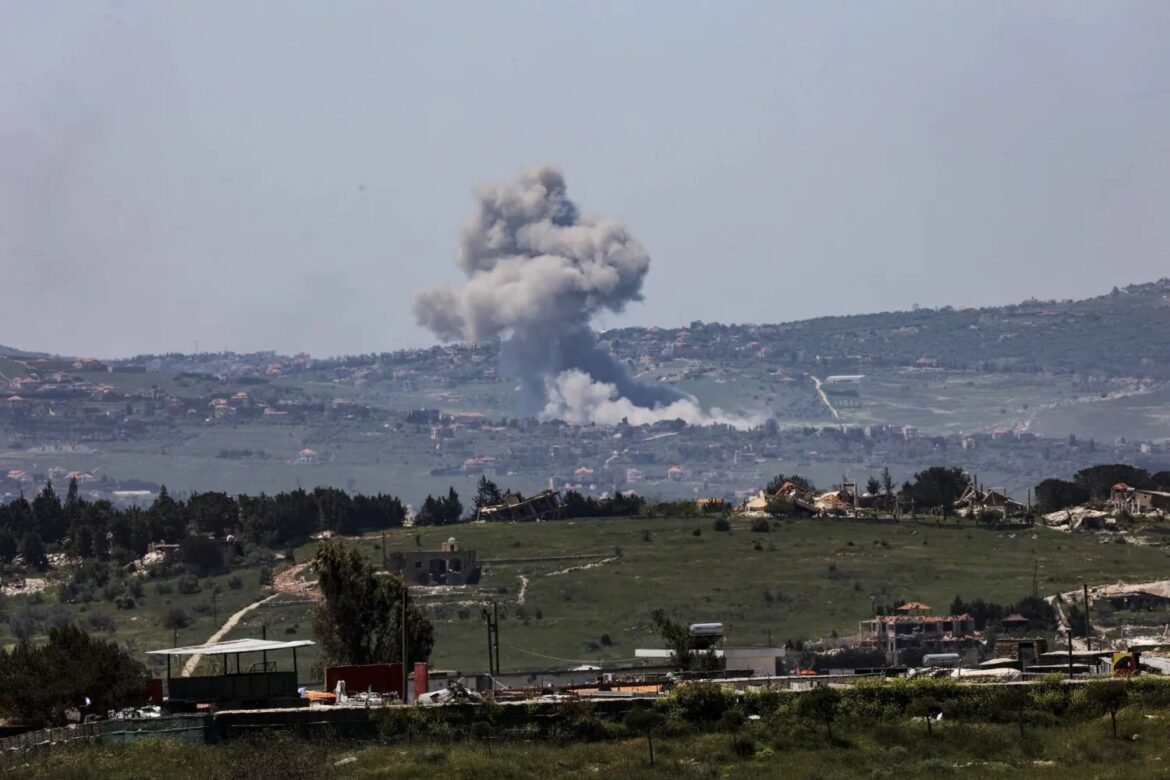 Explosion and large smoke plume rising from a hillside with scattered buildings in the foreground.