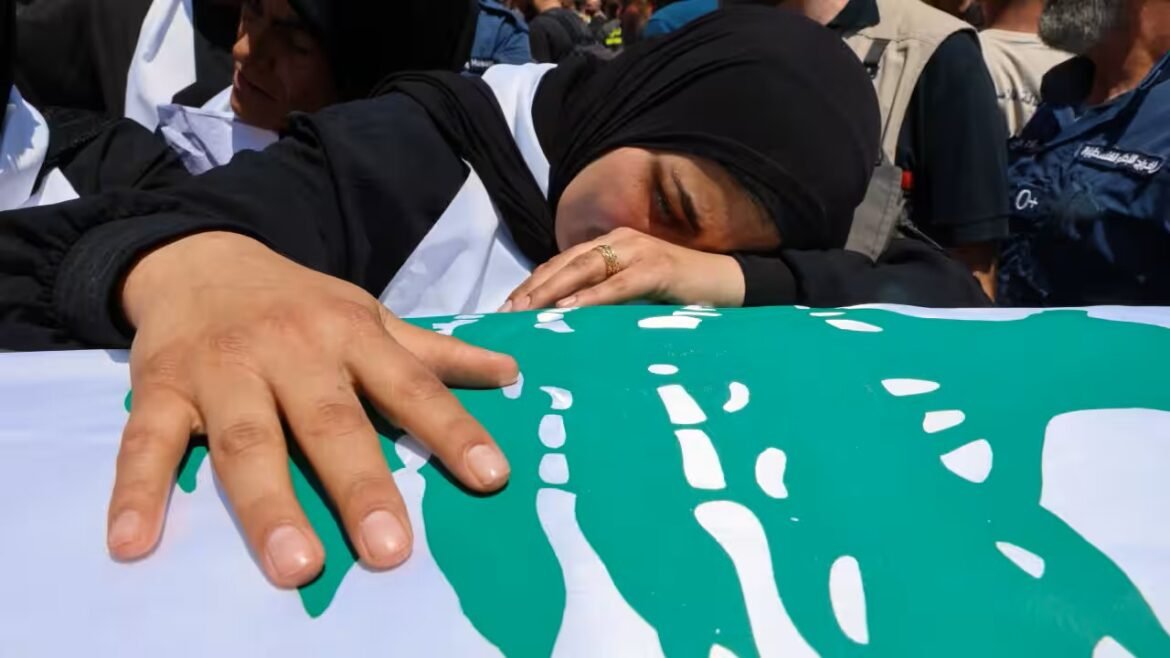 Woman in a black hijab rests her head and hand on a green-and-white banner at a crowded gathering, conveying mourning or solidarity.