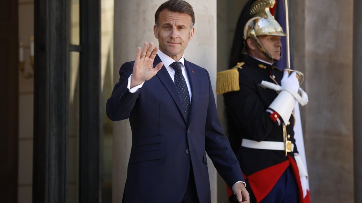 Man in a dark suit waves from a doorway beside a ceremonial guard in ornate uniform.