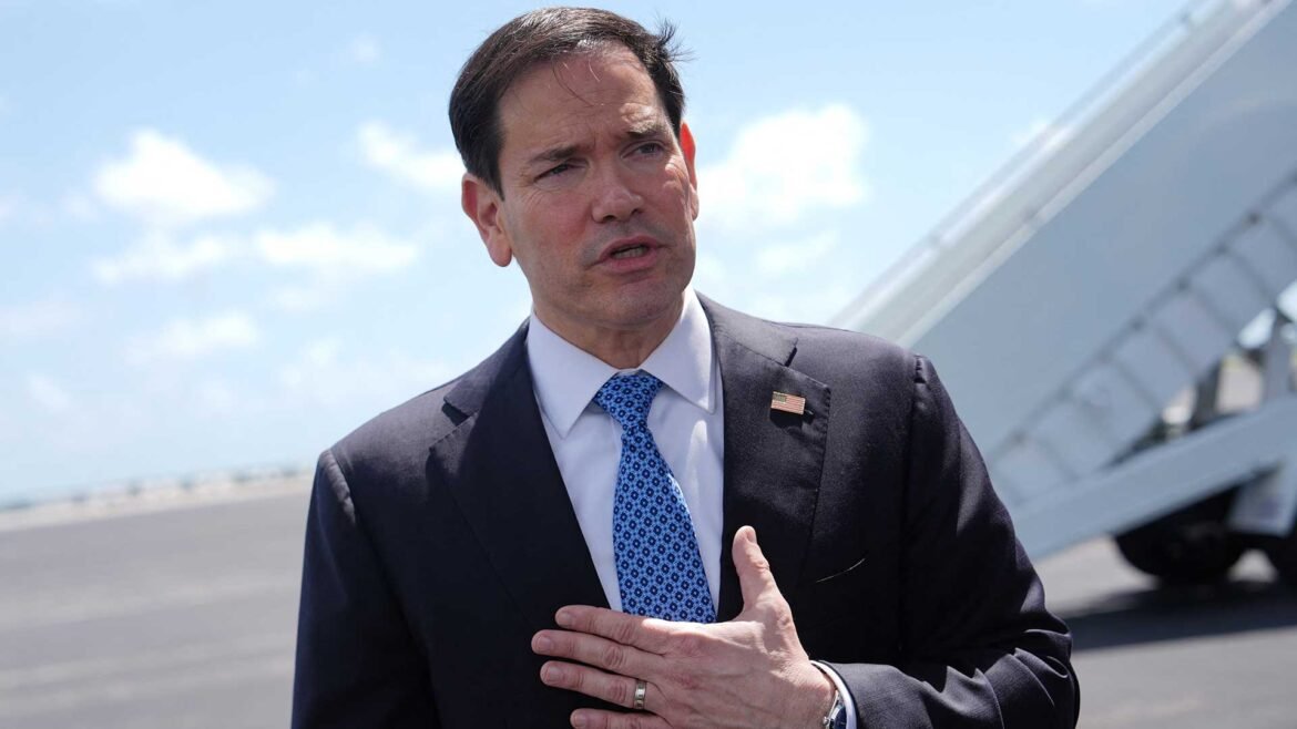 Man in a dark suit and blue patterned tie, wearing an American flag lapel pin, speaking outdoors near an airplane wing.