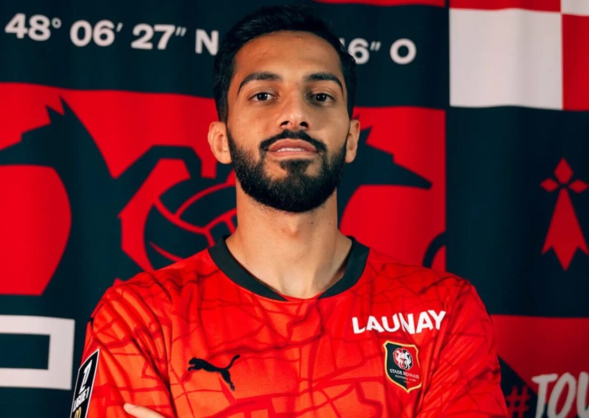 Portrait of a man with a trimmed beard wearing a red sports jersey with 'LAUNAY' and a team crest, standing in front of a red-and-black banner with graphics.