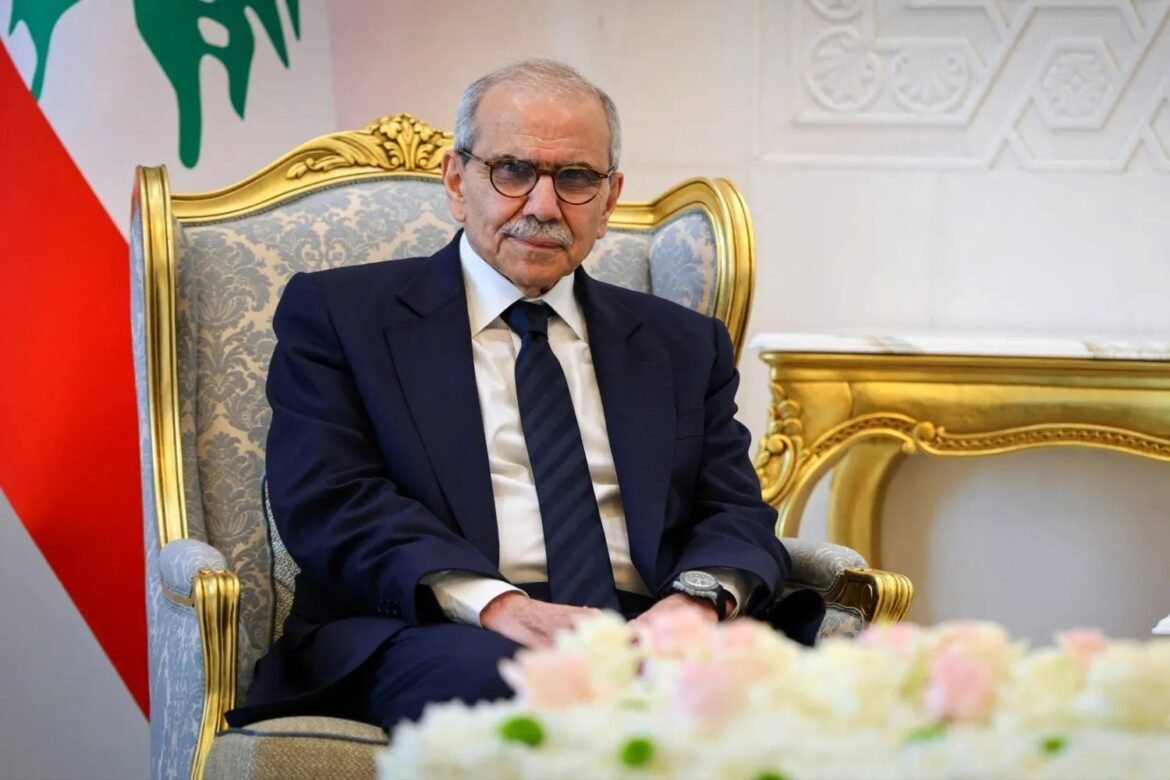 Senior man in a navy suit with glasses sits in an ornate gold-trimmed chair during a formal meeting, with a flag in the background.