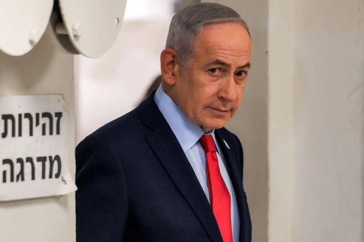 Man in a dark suit and red tie stands in a hallway beside a Hebrew sign, looking to the side.