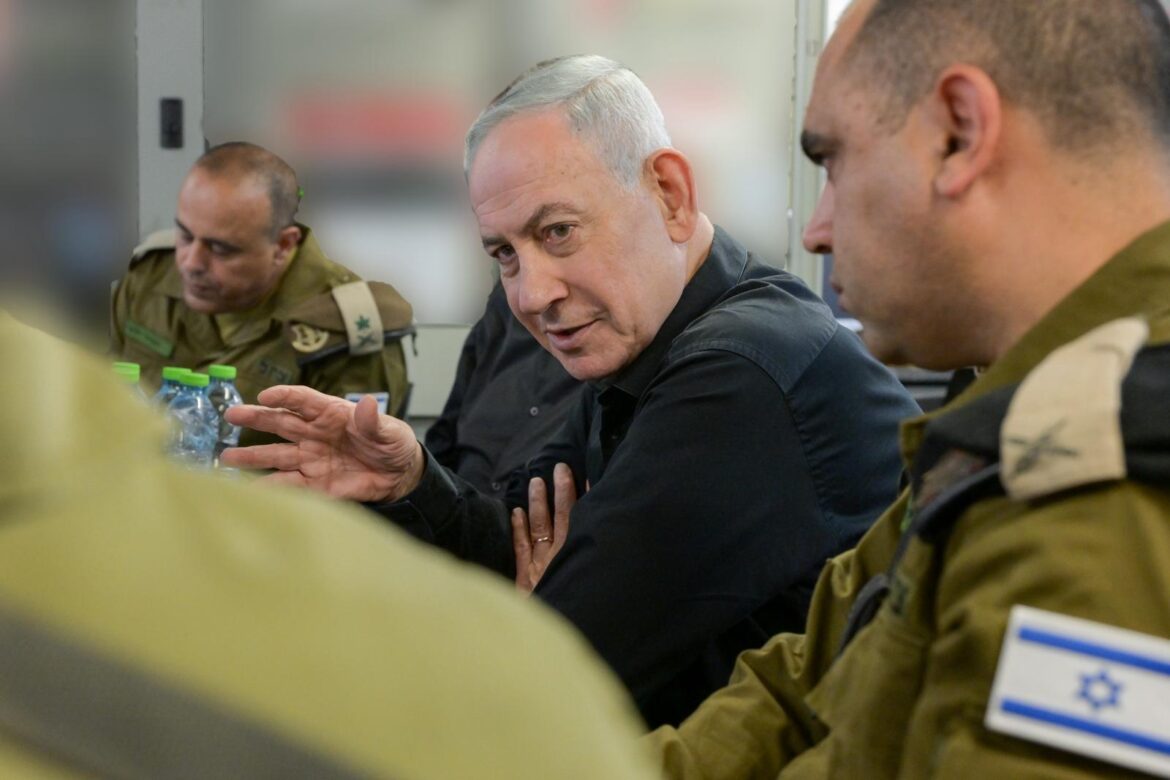 Man with gray hair in a black shirt speaks and gestures while seated with soldiers in tan uniforms around a table, Israeli flag patch visible on one sleeve.