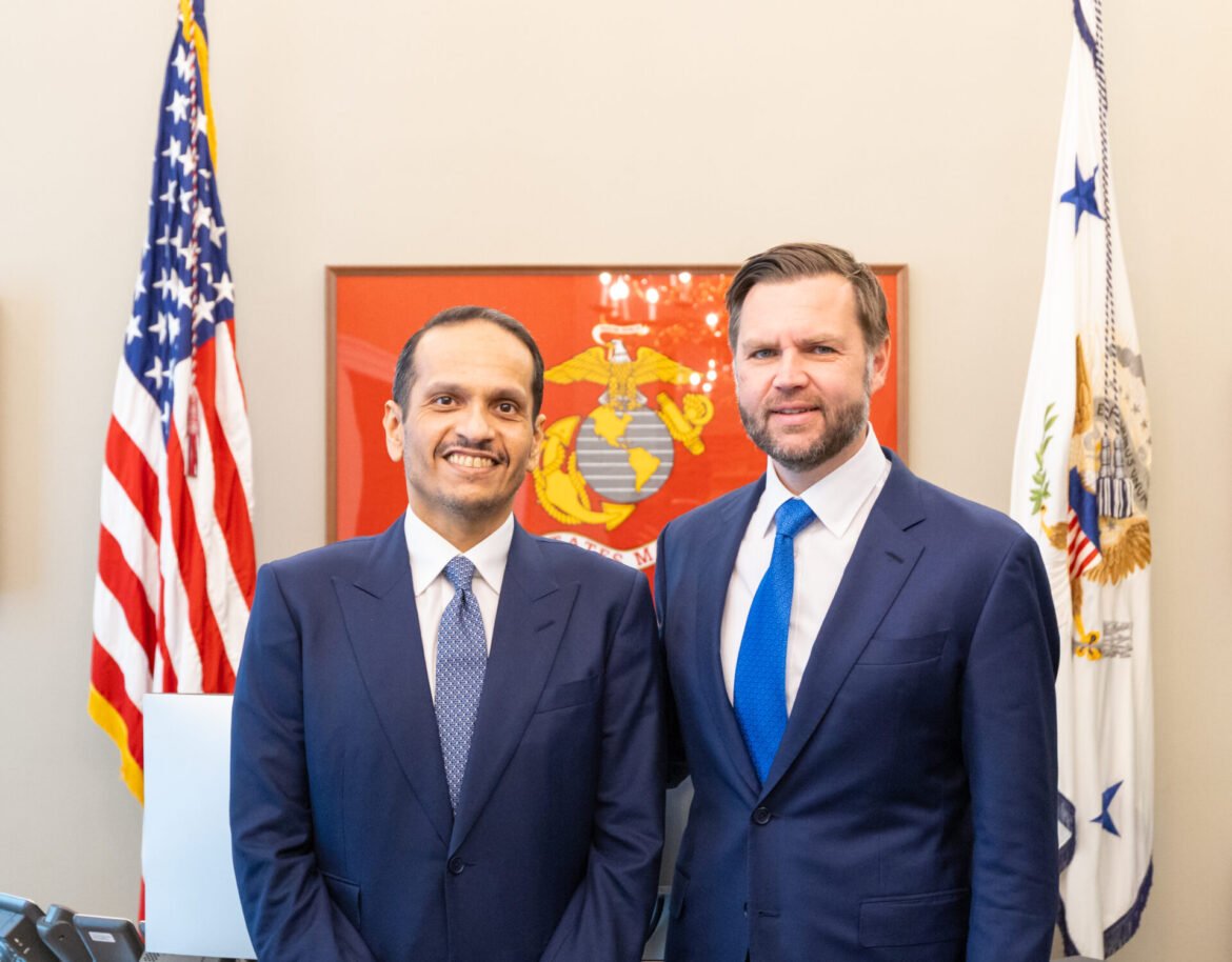 Two men in navy suits stand side by side indoors, smiling, with the American flag and another flag behind them.