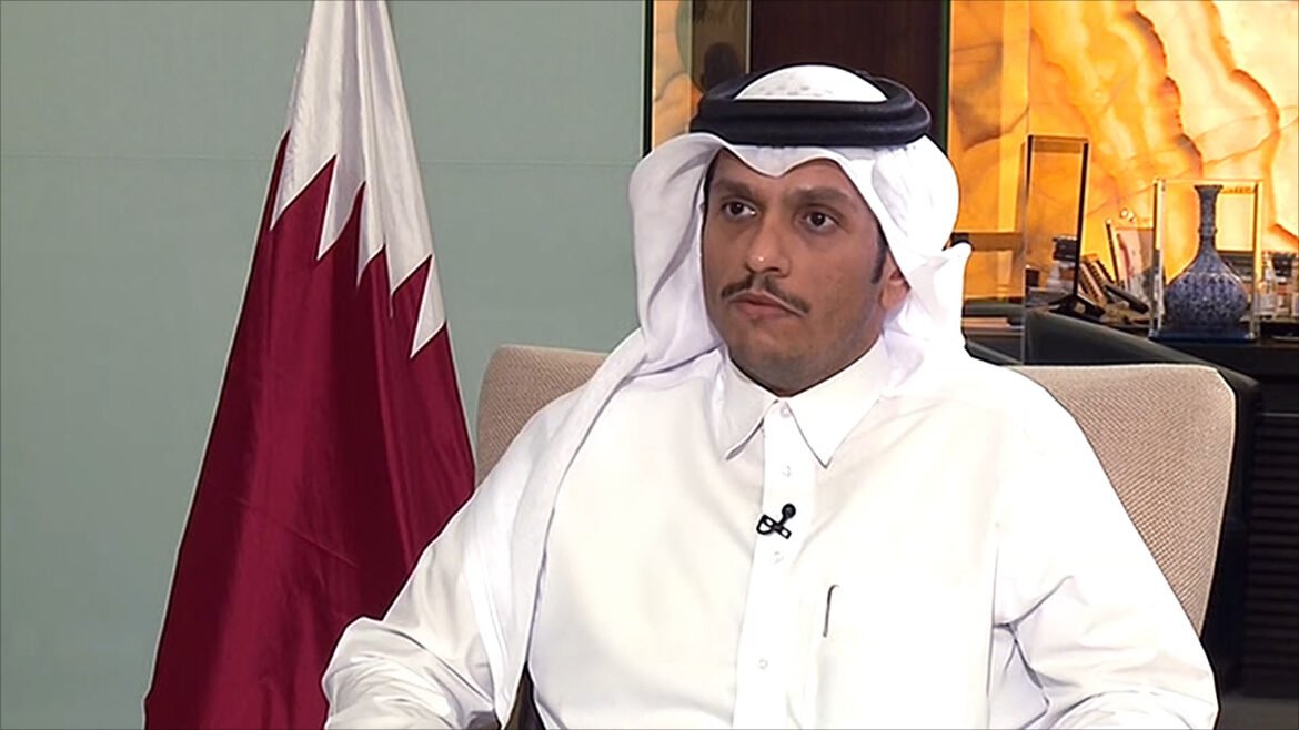 Man in a white thobe and ghutra with a mic clipped to his shirt, seated indoors with a Qatari flag in the background.