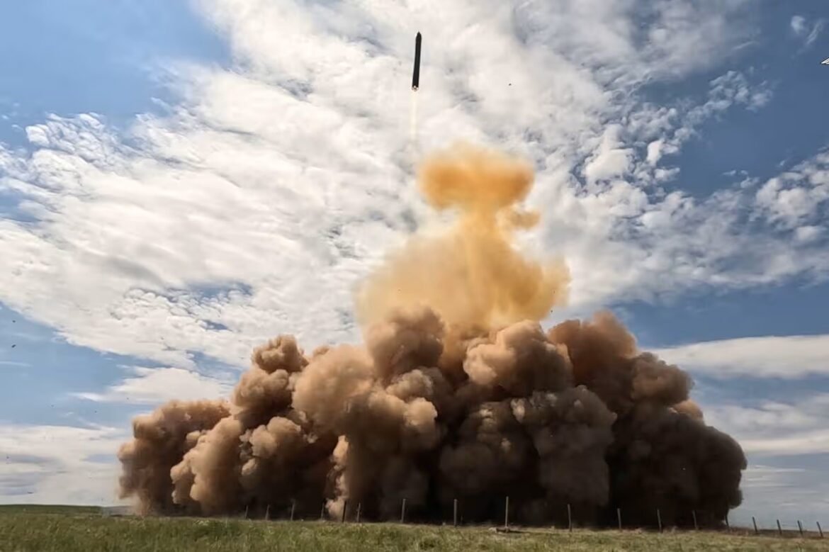 Rocket launches skyward from a grassy field, leaving a massive brown smoke plume as it ascends into a blue sky with scattered clouds.