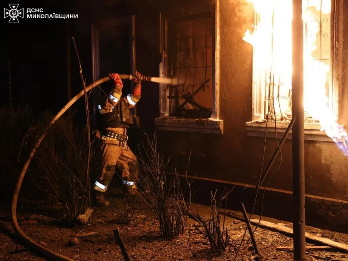 Firefighter in turnout gear spraying water at a burning building at night, flames bursting from a window on the right side, emergency services logo visible in the corner.