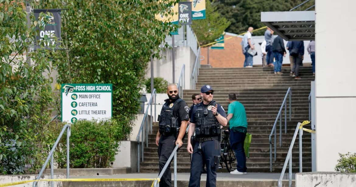 Two uniformed police officers in tactical gear stand at a school entrance, with yellow caution tape across the steps in front of them.