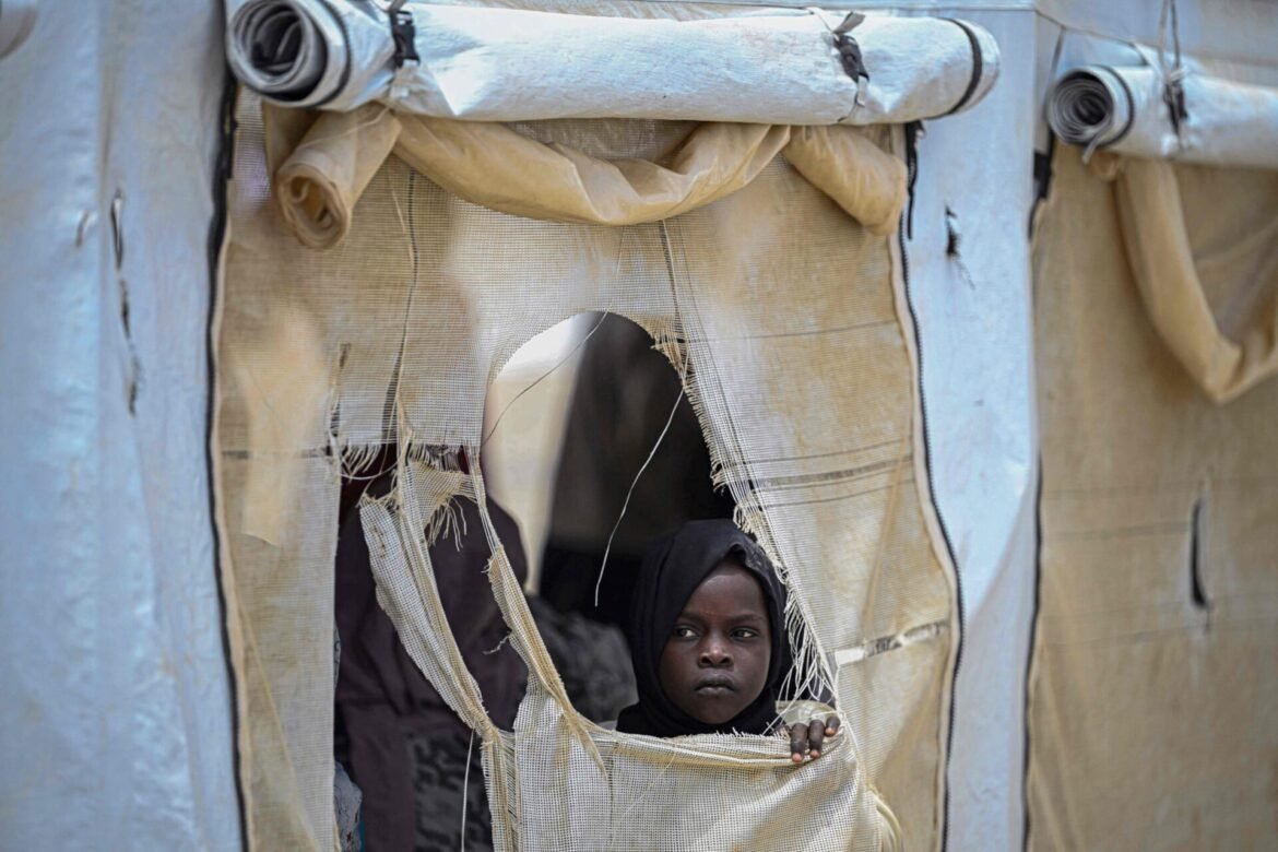 A young girl with a dark headscarf peers through a large torn hole in a beige fabric wall of a shelter or tent Ori, looking outward with a serious expression.