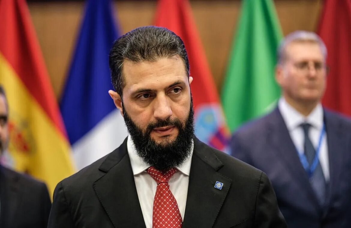 Bearded man in a black suit and red tie at a diplomatic event, with colorful flags in the background.