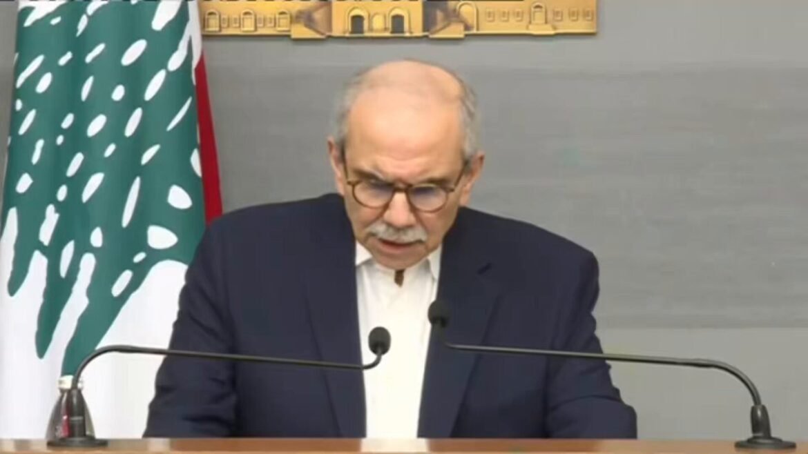 A man in a dark suit and glasses speaks from a podium with two microphones, Lebanese flag visible to the left.