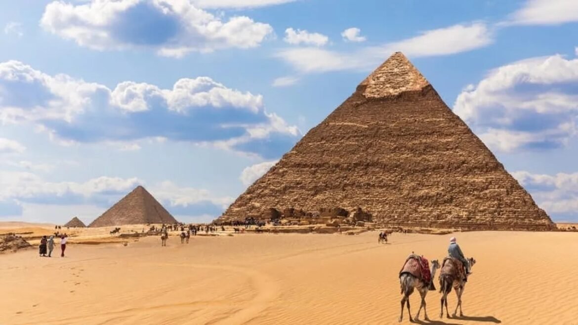 Camel riders approach the Great Pyramid of Giza on a sunny desert day.