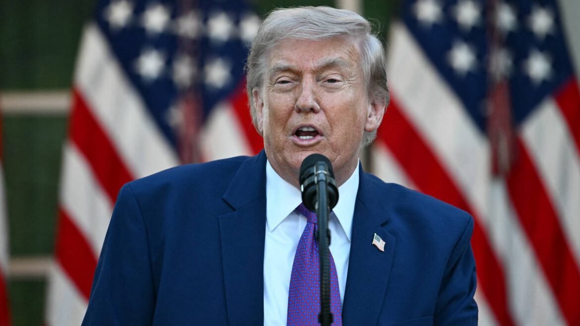 Man in a blue suit and red tie speaks into a microphone with American flags in the background behind him.