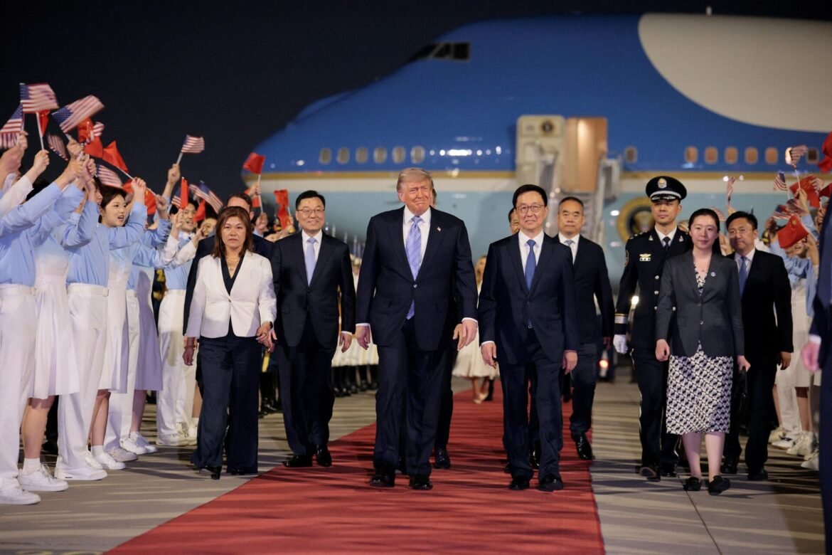Group of world leaders walking on a red carpet at night, supporters waving American flags with a large blue airplane in the background