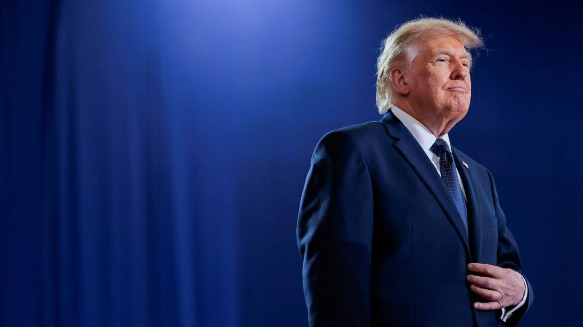 Man in a dark navy suit and tie stands on a stage, looking toward the right with a blue backdrop behind him.