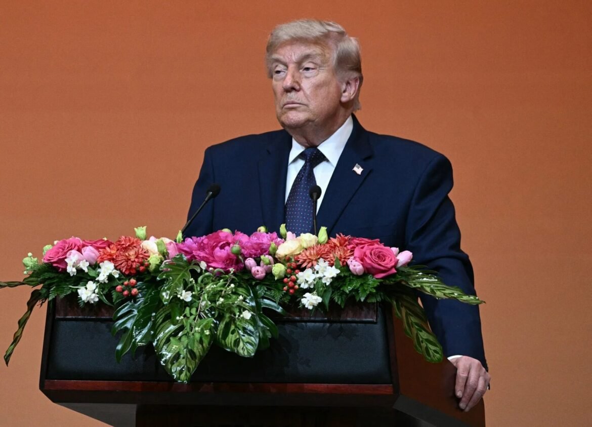 Man in a dark suit and tie stands at a podium with a large flower arrangement, speaking at a formal event against an orange backdrop.