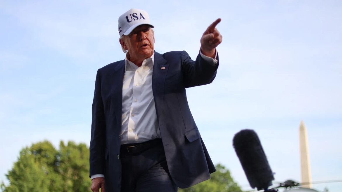Man in a dark suit and white USA cap points forward at an outdoor rally, with a microphone in the foreground.