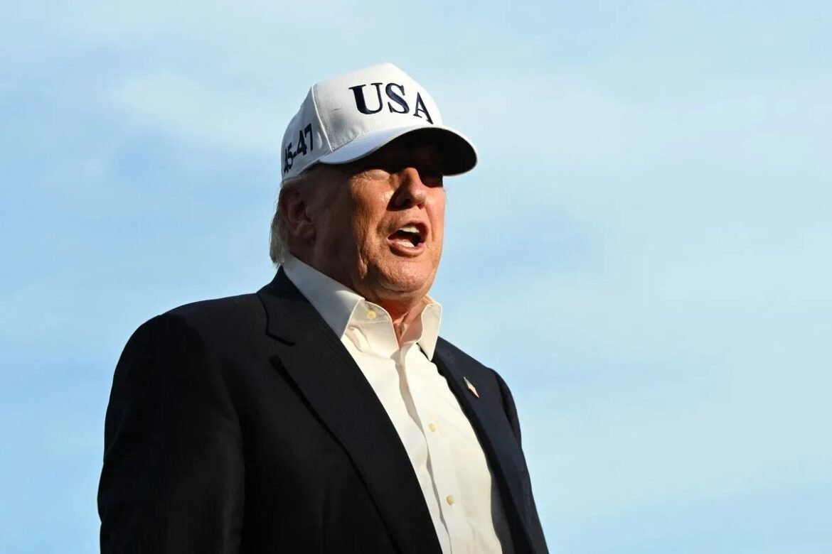 Man in a dark suit and white shirt wearing a white cap with 'USA', speaking outdoors against a blue sky.